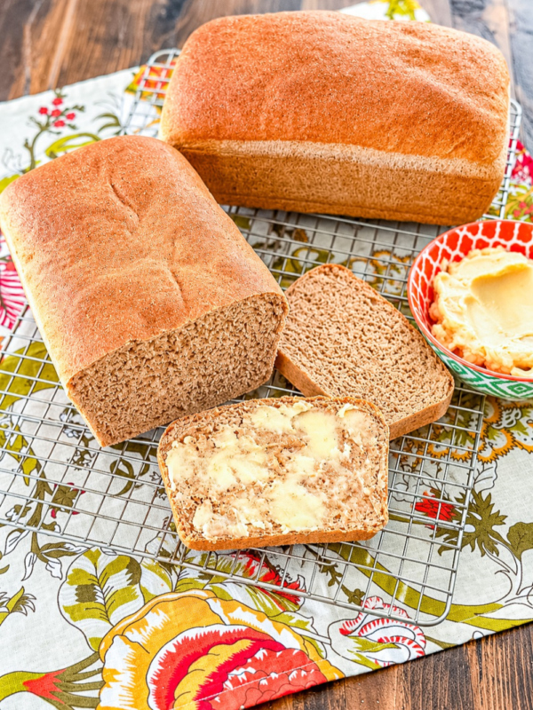 Slices of one hour wheat bread, one topped with honey butter, beside two freshly baked loaves