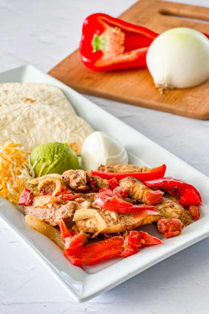 Close up of Oven Baked Chicken Fajitas on a white platter with tortillas, cheese, guacamole, and sour cream