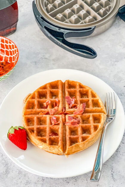 cinnamon waffle sprinkled with bacon on a white plate with a strawberry and fork