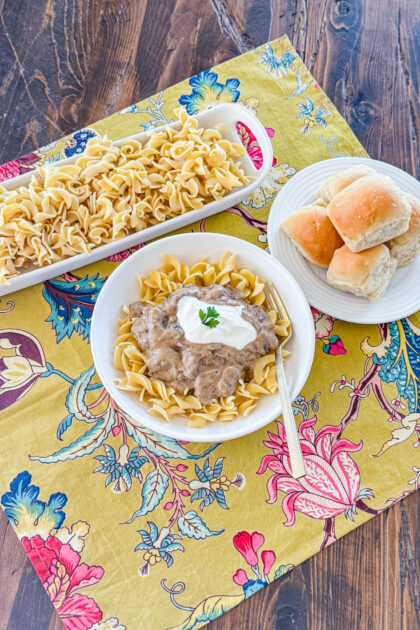 bowl of egg noodles tops with mushroom beef stroganoff sauce, a dollop of sour cream, and chopped green onion with a dish full of noodles and a plate of rolls on the side