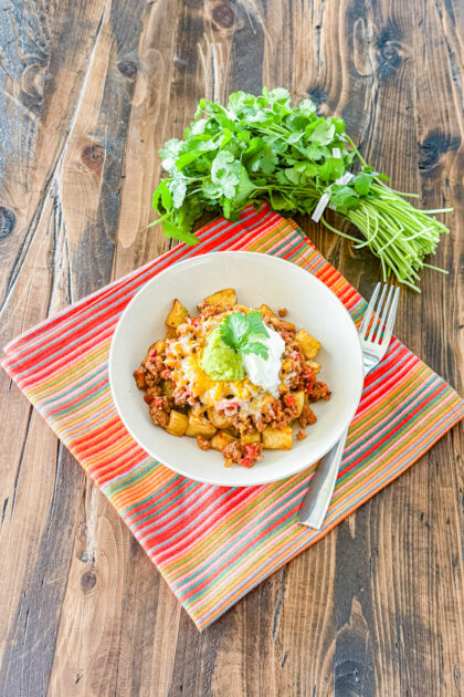 a bowl of roasted potatoes topped with taco meat, shredded cheese, a spoonful of guacamole and sour cream with a cilantro leaf on top