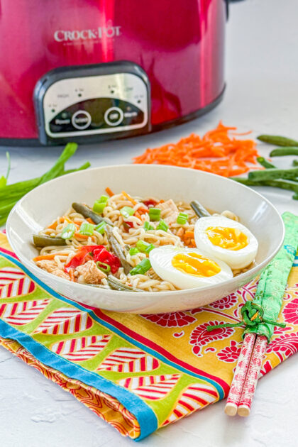bowl of ramen noodle soup with vegetables and soft bpoiled egg next to a slow cooker