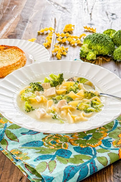 bowl of creamy chicken broccoli soup in a bowl with gaarlic bread, noodles, and broccoli in the background