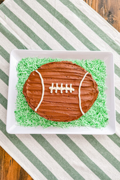 football shaped chocolate cake on a white tray surrounded with green colored shredded coconut