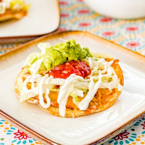 Close-up of an oven-baked tostada with refried beans, shredded cheese, lettuce, sour cream, guacamole, and salsa