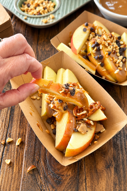 Hand holding a caramel apple slice topped with toffee and pretzel pieces