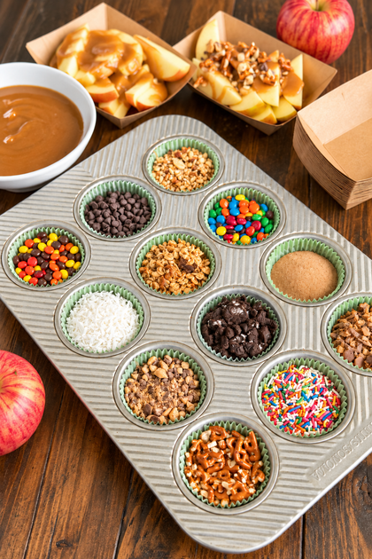 Muffin tin filled with caramel apple toppings including chocolate chips, candy, nuts, coconut, sprinkles, and pretzels