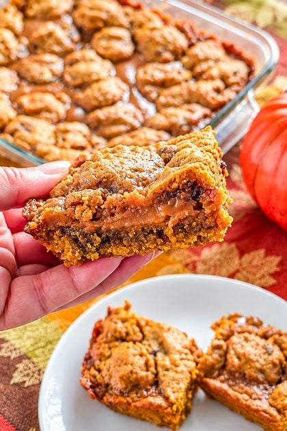 Hand holding a gooey pumpkin caramel bar with the baking dish and fall decor in the background