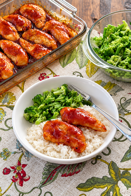 Sweet sticky chicken tenders over white rice with broccoli, with the baking dish of chicken in the background